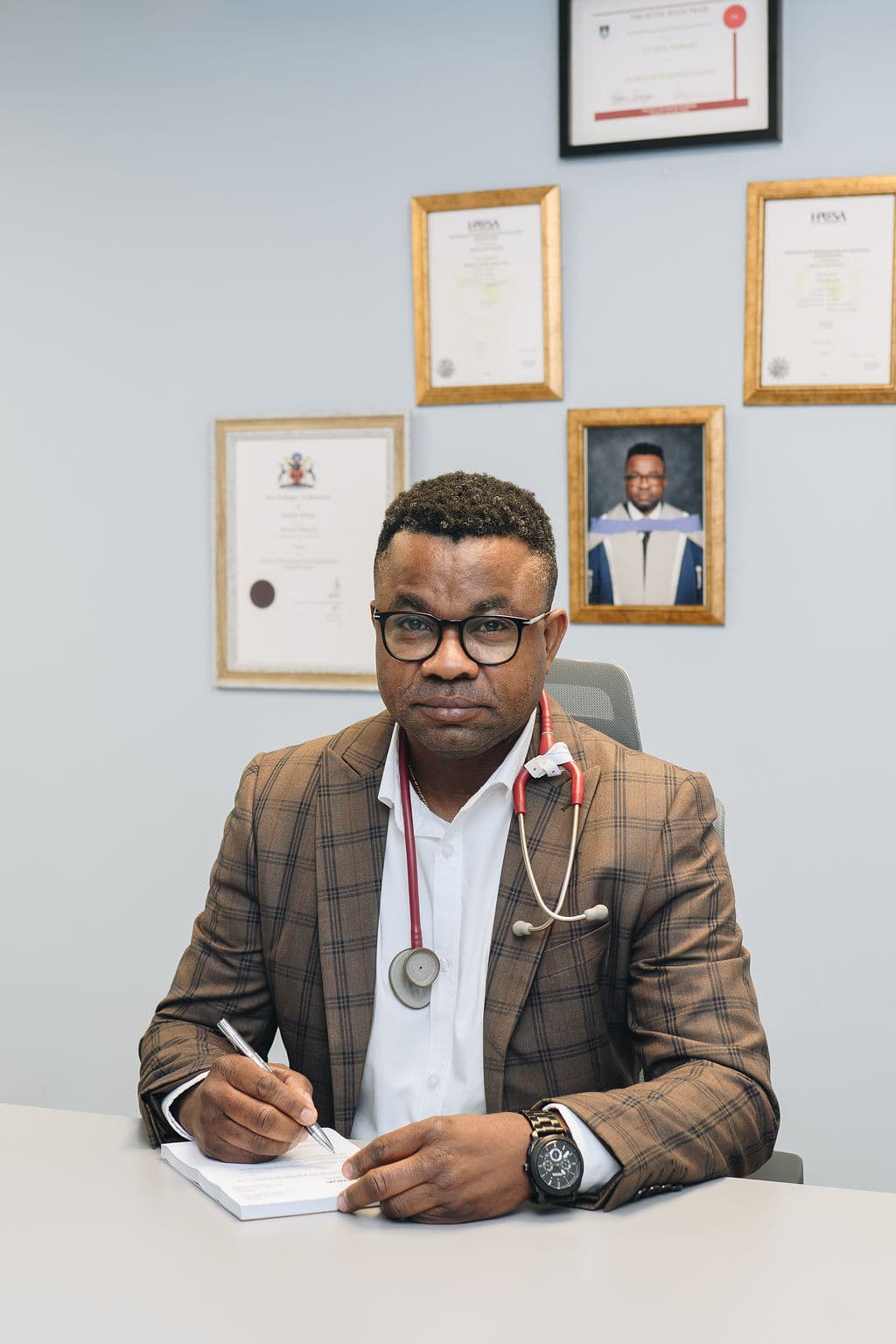 Dr Malonda Makanda, specialist obstetrician and gynaecologist, seated at his consulting-room desk in a brown check blazer with a stethoscope around his neck, writing notes, with his framed medical qualifications and HPCSA registration certificates on the wall behind