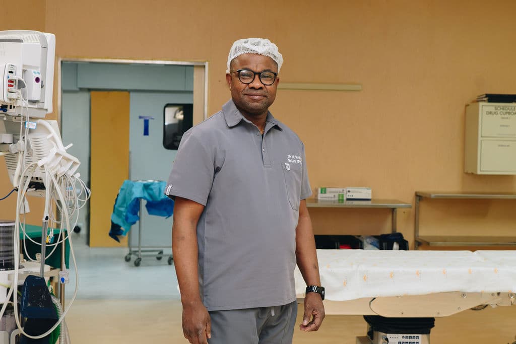 Dr Makanda smiling in a theatre cap and grey scrubs embroidered with his name, standing in the operating theatre at Melomed Private Hospital, Mitchells Plain